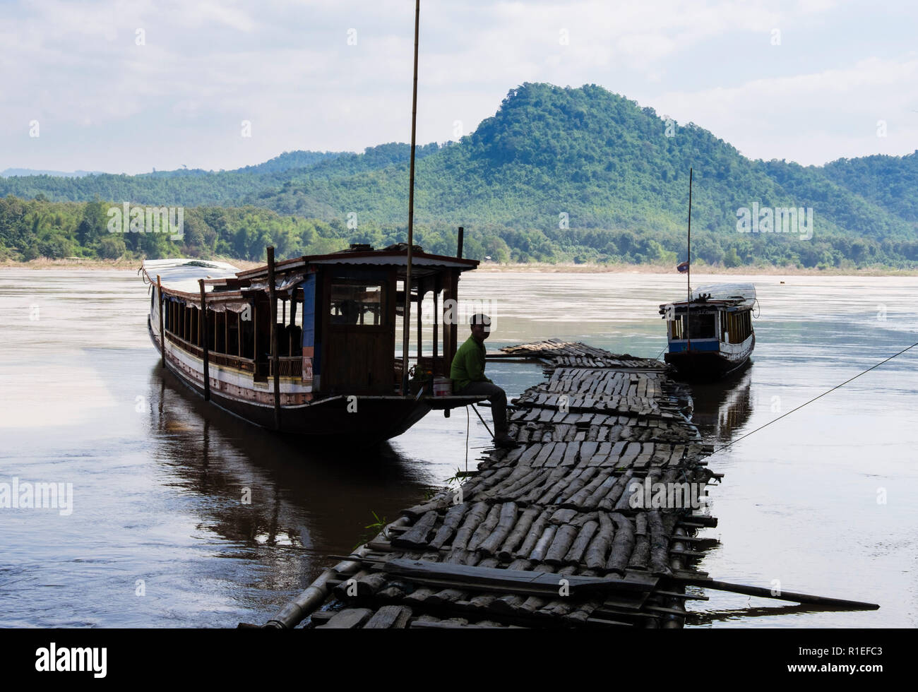 Boatman waits by tourist boats moored by flimsy wooden jetty at Tham Ting Caves on Mekong River. Pak Ou, Luang Prabang province, Laos, Asia Stock Photo