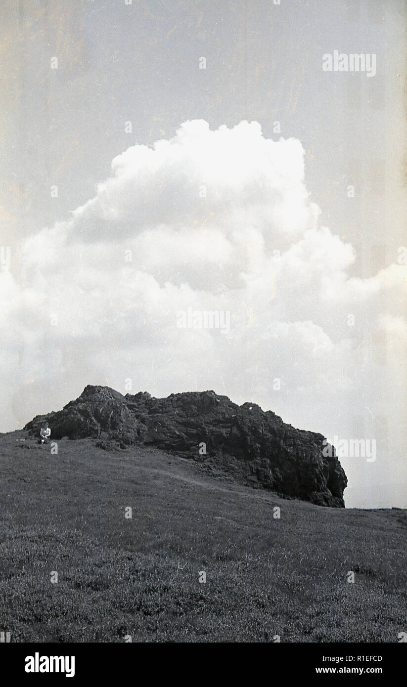 1950s, historical, Exmoor and a view from a distance of a tor or craig, a rocky peak that juts out on the landscape. A female walker can be seen sitting by by the rocks. Stock Photo