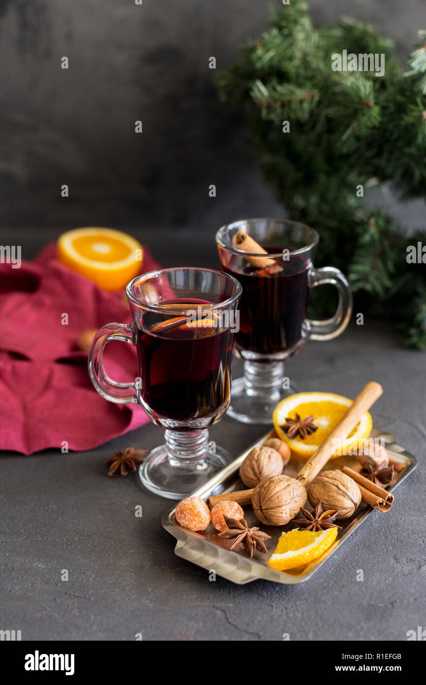 Winter christmas composition. Red mulled wine in glasses at black background. Fir wreath, tray with orange, cinnamon, nuts, cone and spices near. Stock Photo
