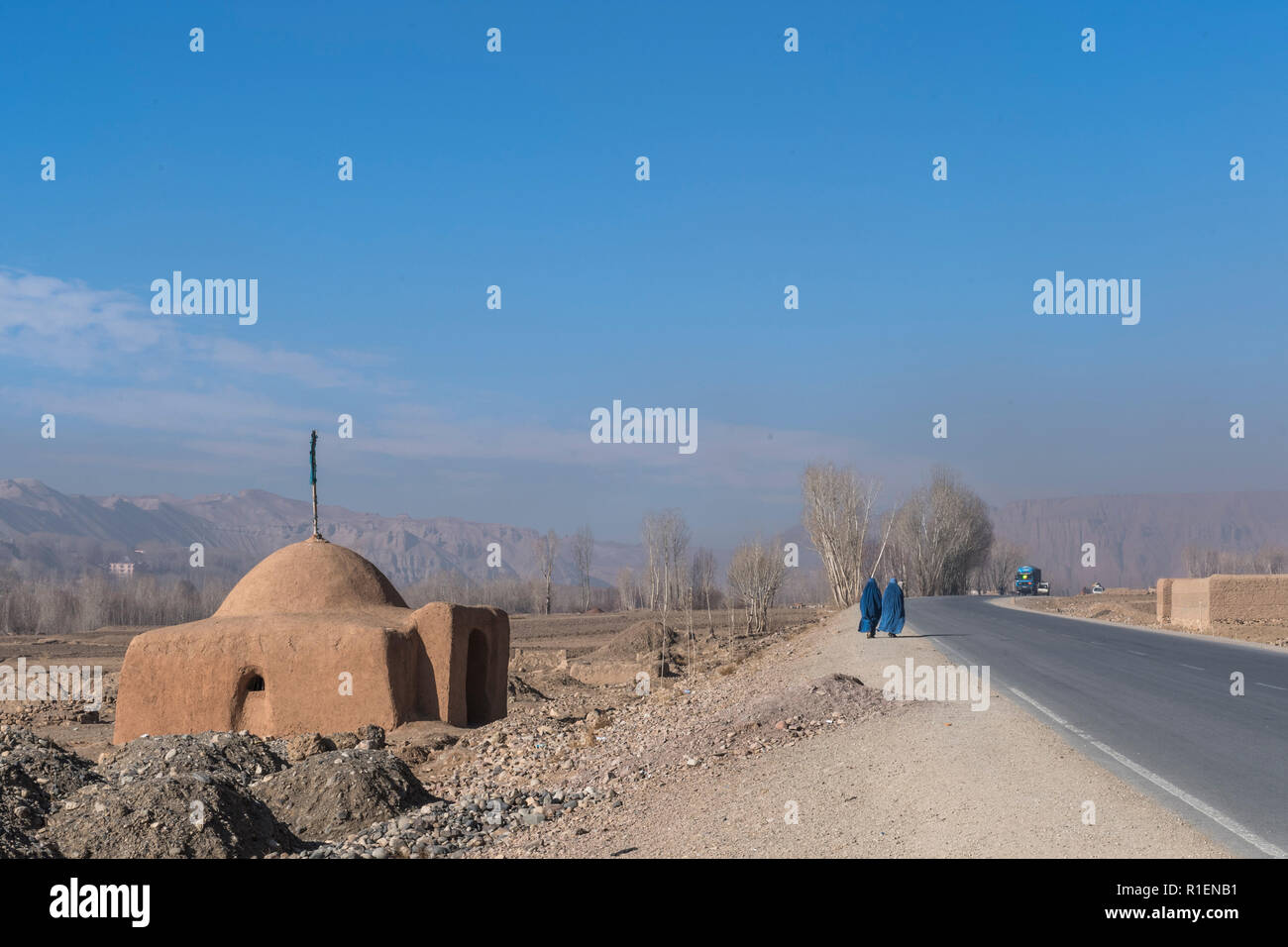 2 Ladies Wearing Blue Burqa Walking On Road Entering The Bamyan Valley With A Shrine And Mountains In The Background, Bamyan Province, Afghanistan Stock Photo