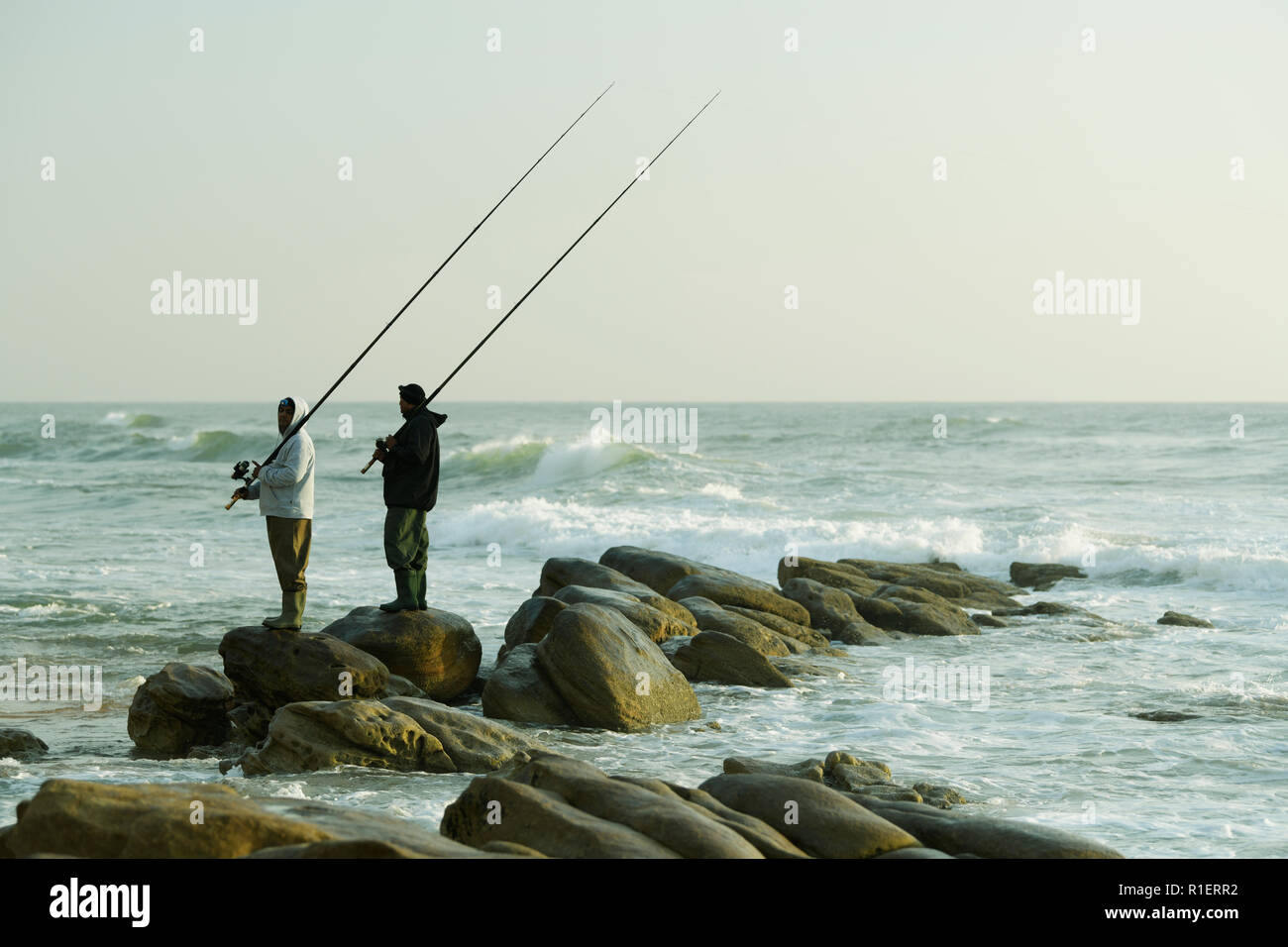 Durban, KwaZulu-Natal, two adult male fishermen in waterproof clothing standing on wet rocks with fishing rods to catch fish at Umhlanga Rocks beach Stock Photo