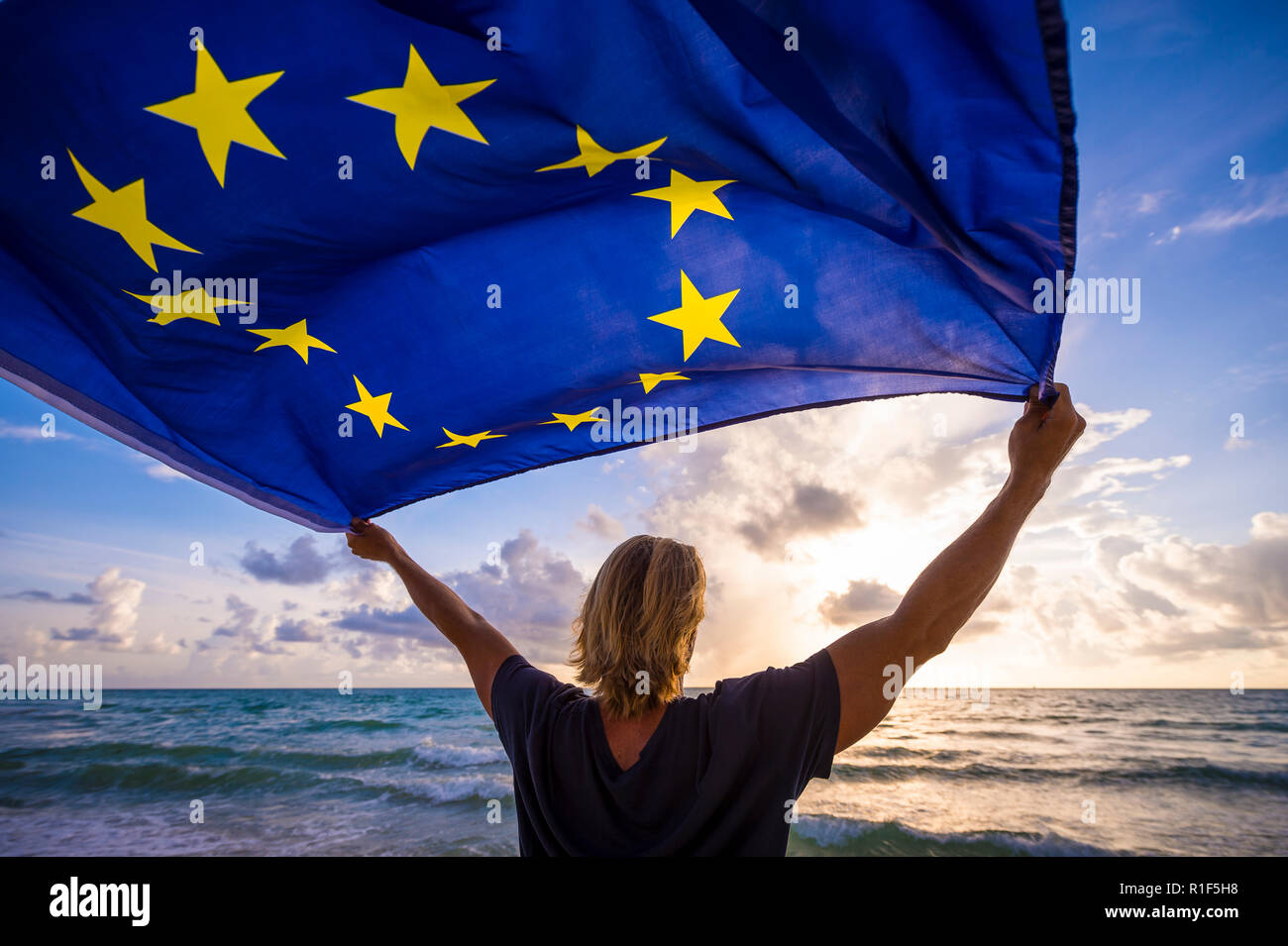 Man with blond hair holding EU European Union flag waving in the wind of a Mediterranean beach scene Stock Photo