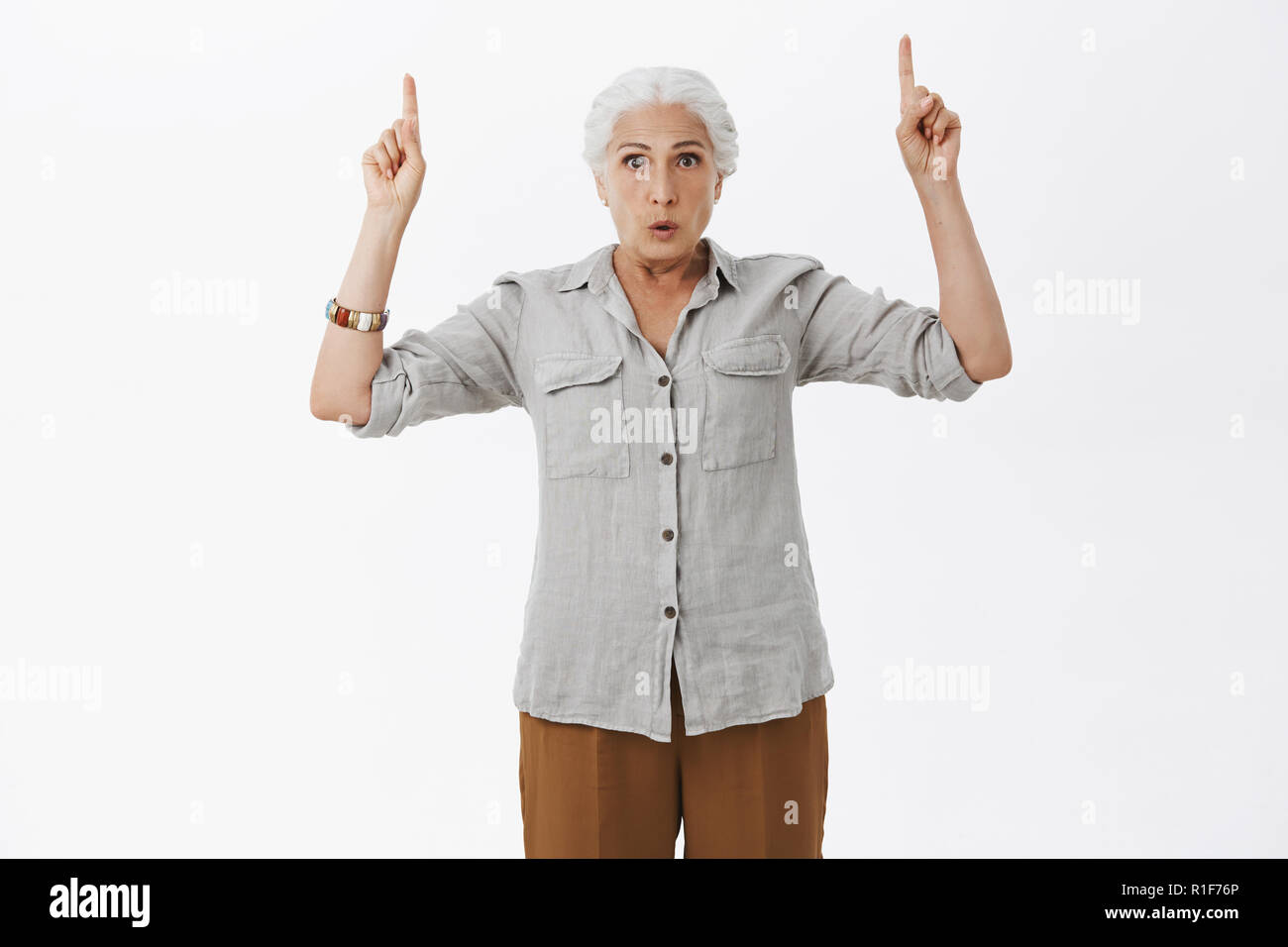 Indoro shot of worried questioned granny with grey hair in casual shirt raising hands pointing up folding lips and gazing strict at camera with worrie Stock Photo