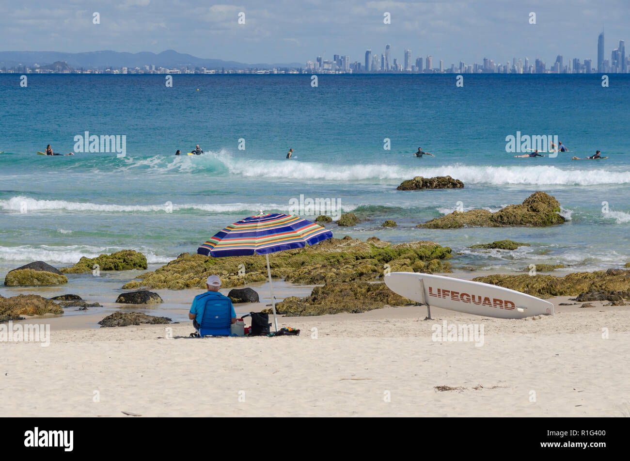 Lifeguard sits on beach in the shade of a umbrella watching surfers, with city skyline of Surf Paradise in background. Stock Photo