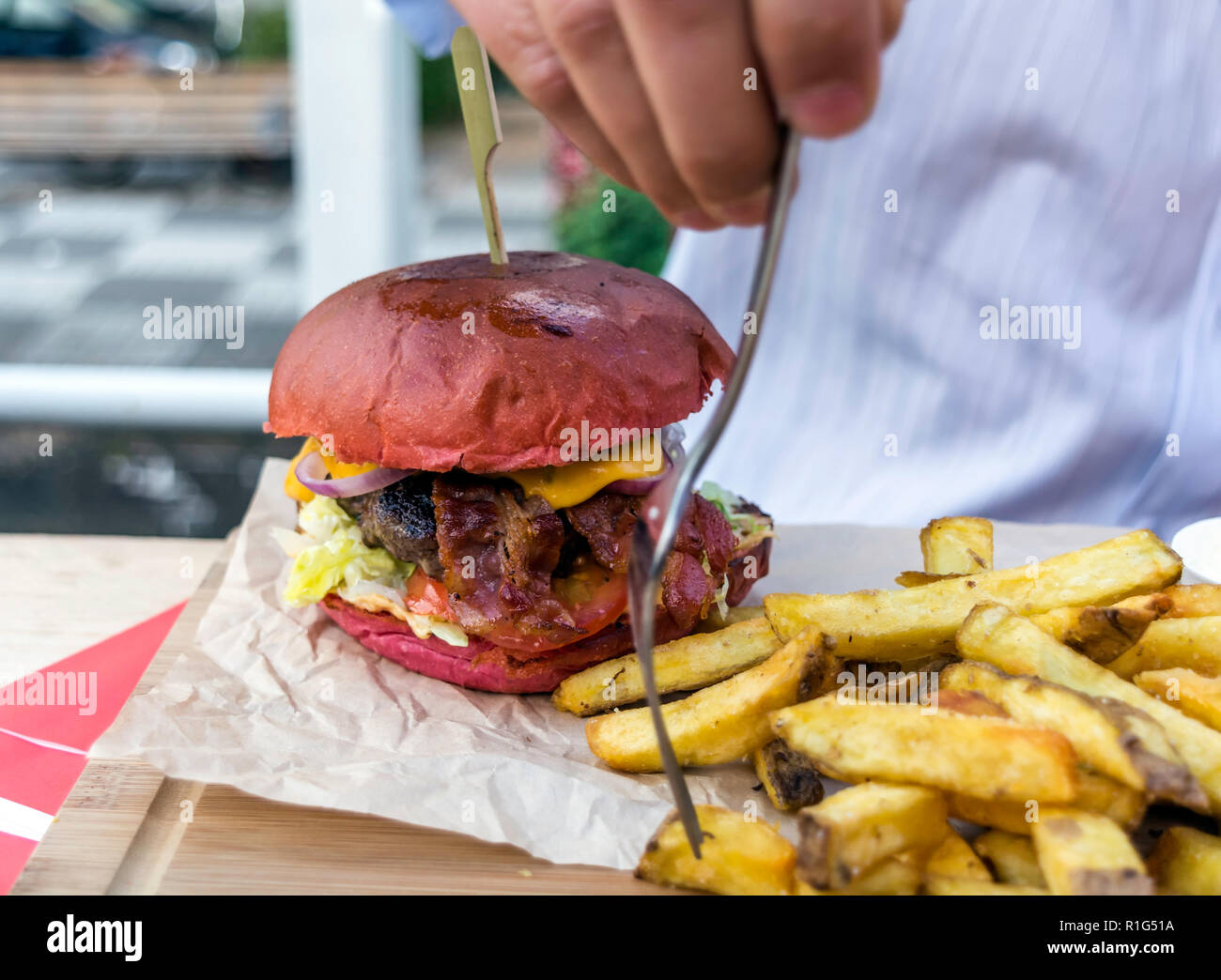 Giant juicy burger with various toppings and tasty meat served with large homemade fries on a wooden plate. Male hand taking fried potato with fork. Stock Photo