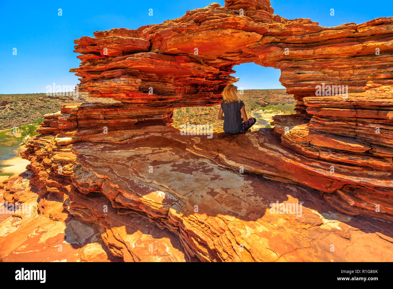 Blonde caucasian woman sitting inside the iconic rock arch in red sandstone of Nature's Window, Kalbarri National Park. Caucasian young girl in Western Australia looking australian outback landscape. Stock Photo