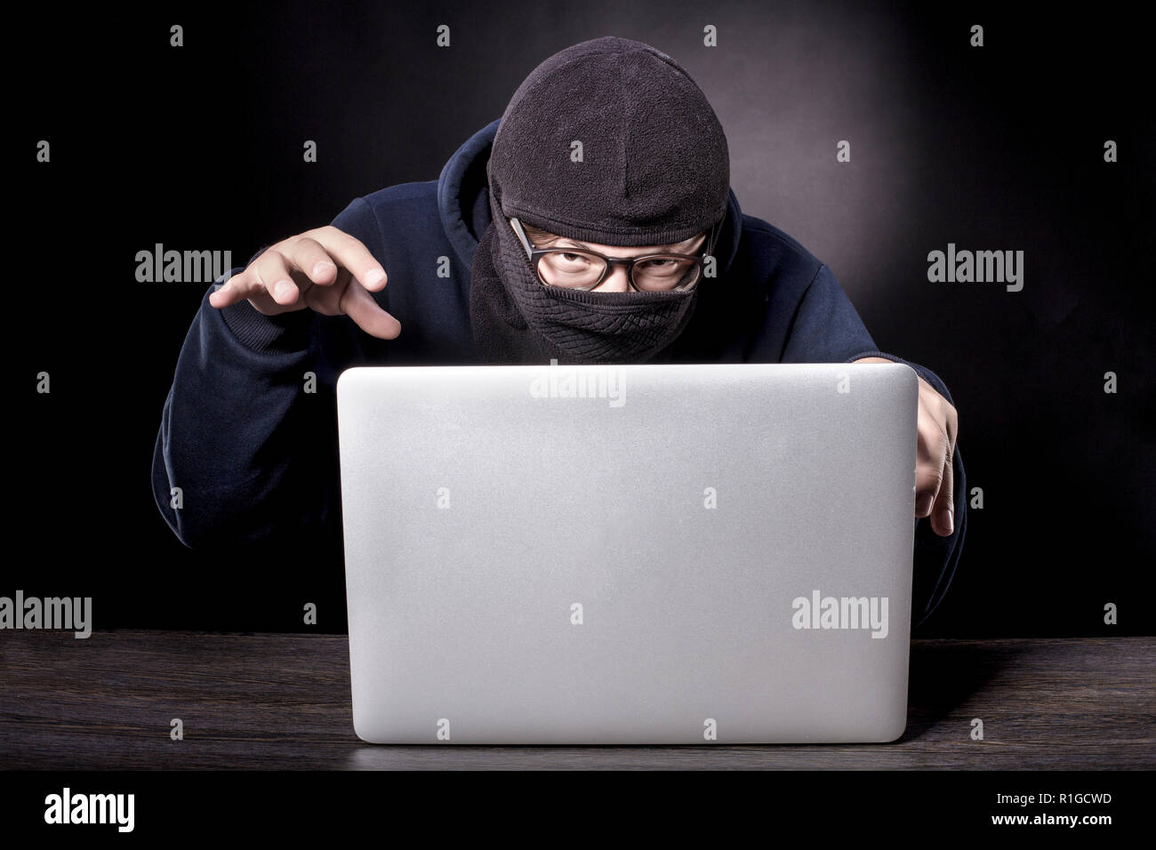 the cunning hacker in balaclava is sitting at the laptop and slyly looking at the camera Stock Photo