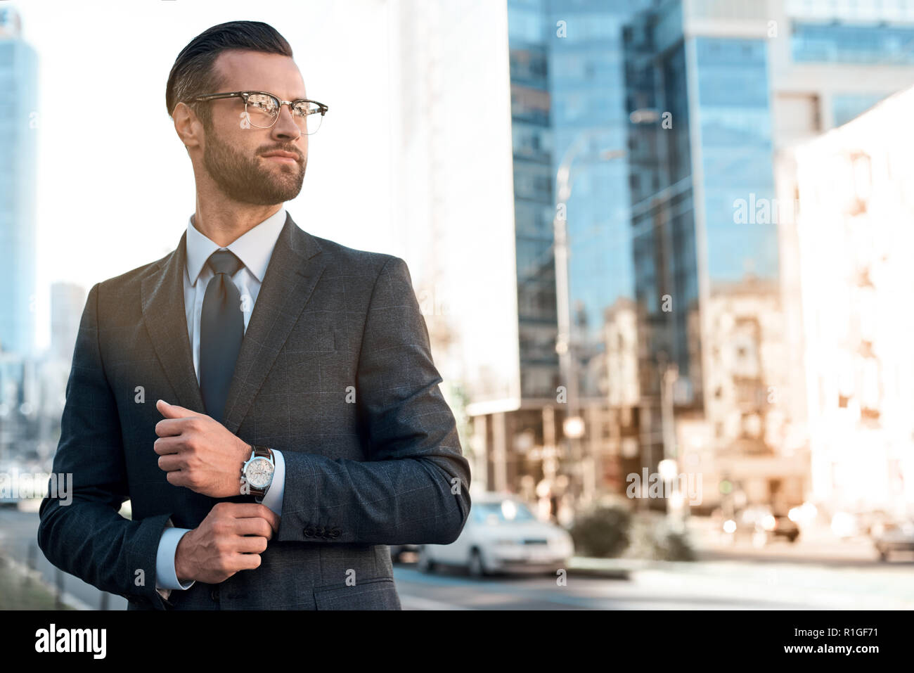 Close up profile portrait of a successful young bearded guy in suit and glasses. So stylish and nerdy. Outdoors on a sunny street, fixing his cuffs Stock Photo