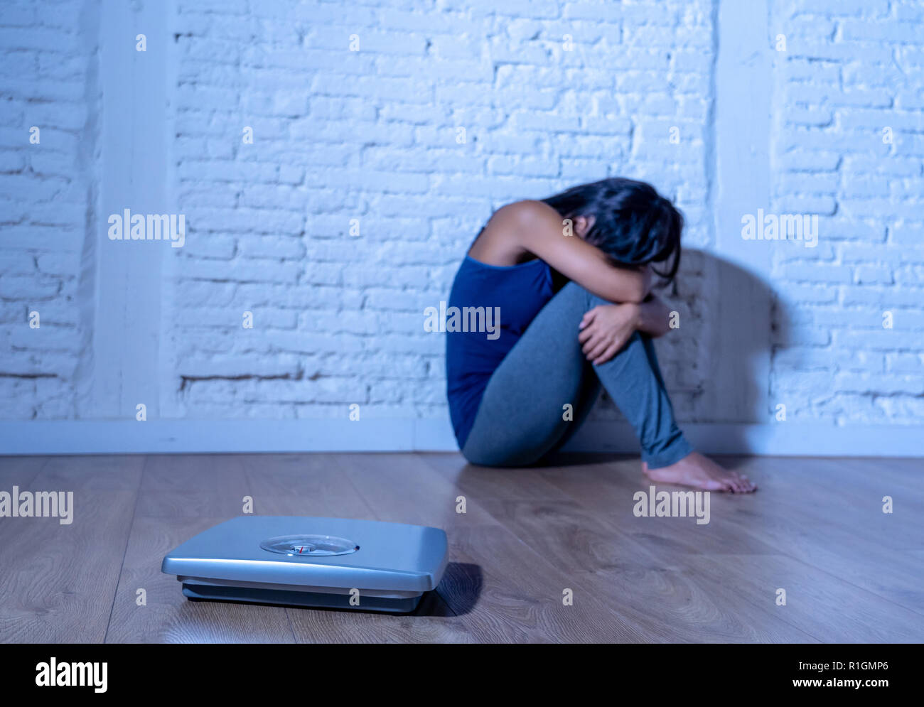 Young anorexic teenager woman sitting alone on ground looking at the scale worried and depressed in dieting and eating disorder concept Stock Photo