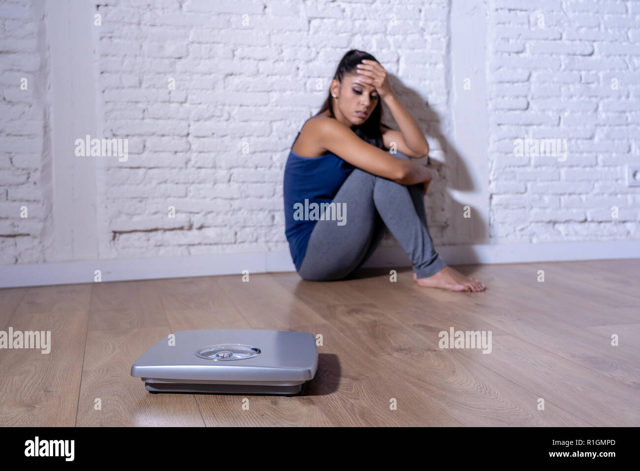 Young anorexic teenager woman sitting alone on ground looking at the scale worried and depressed in dieting and eating disorder concept Stock Photo