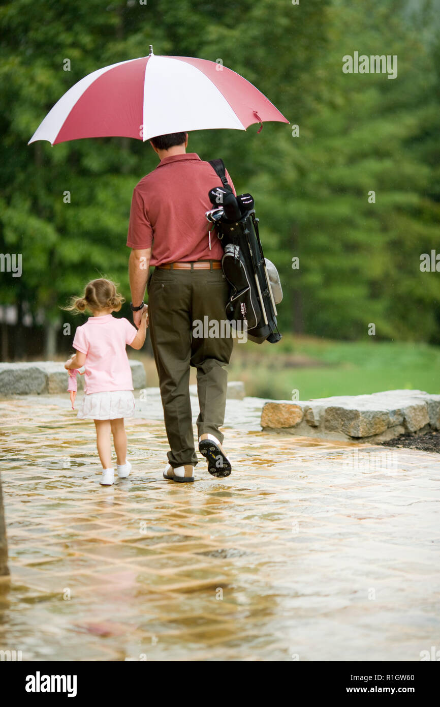 Young girl holding her mid-adult father's hand as the walk with a golf bag and an umbrella. Stock Photo