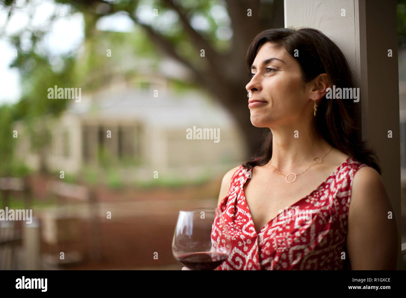 Contented middle-aged woman smiles peacefully as she relaxes with a glass of red wine and looks at the view from her back doorstep. Stock Photo
