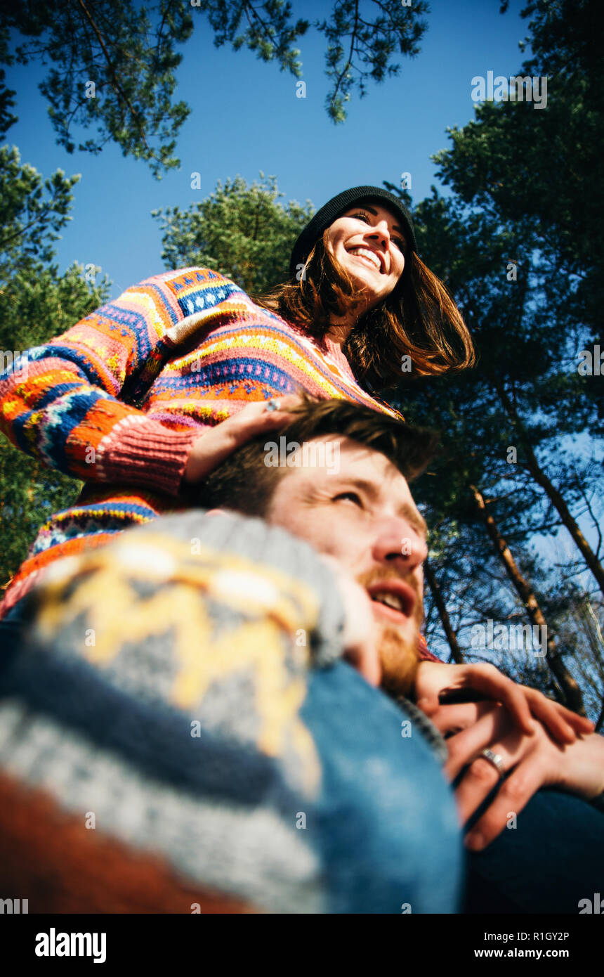 Joyful young girlfriend sits on boyfriend shoulders during walk in forest, happy couple wearing sweaters having fun outdoors Stock Photo