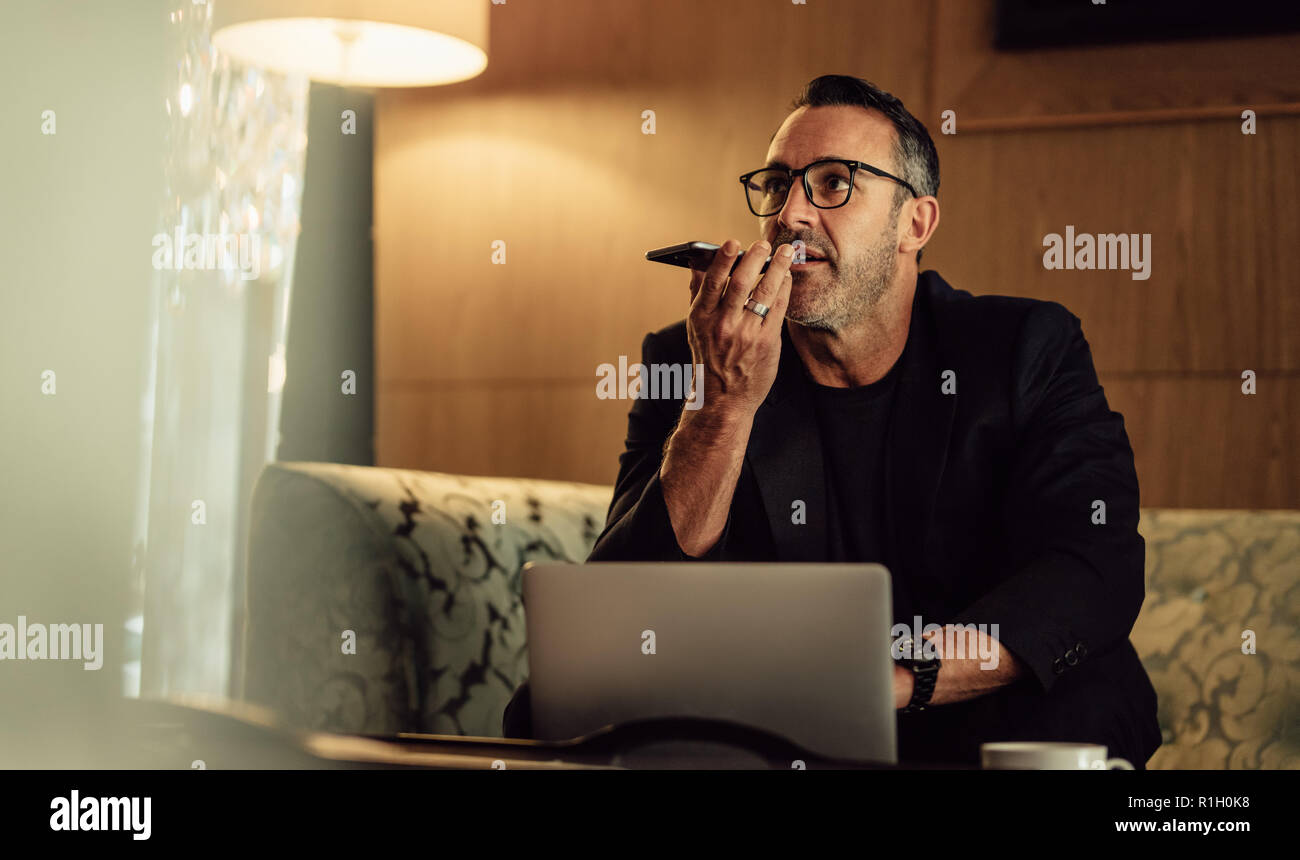 Mature businessman sitting in hotel lobby and talking on mobile phone with laptop in front. Businessman on trip working from hotel lobby. Stock Photo