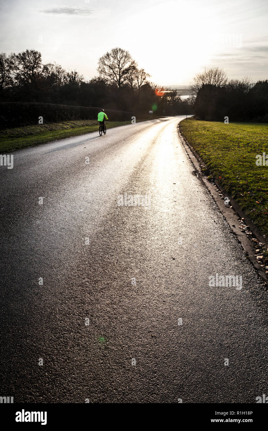 A cyclist in a green/yellow 'hi-viz' jacket, rides along a long damp curve of tarmac road. Stock Photo