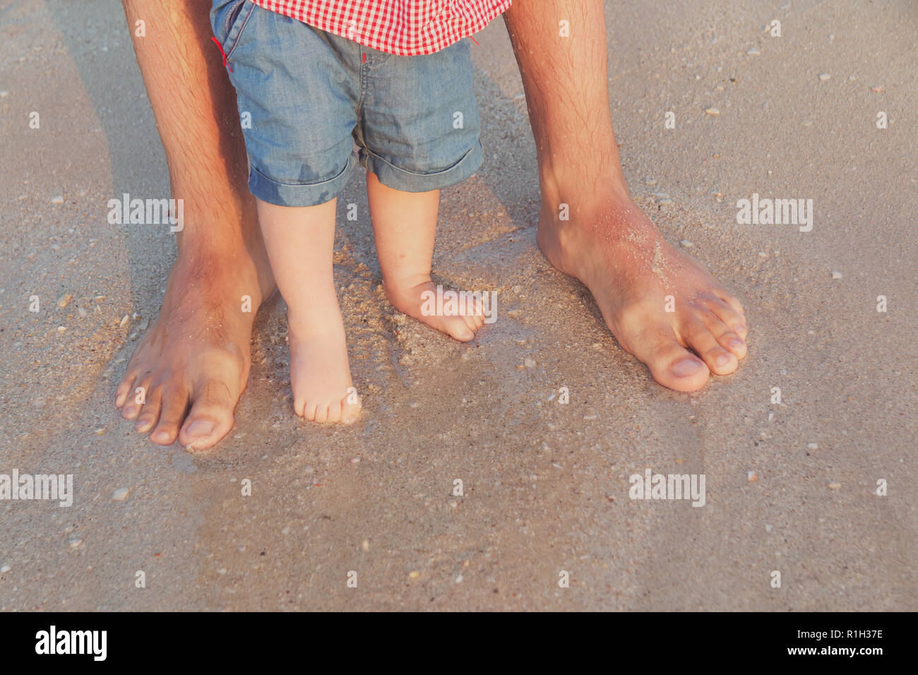 Man and baby feet standing in shallow water waiting for the wave. Bare feet father and his little daughter or son staying in the sand near the sea. Co Stock Photo