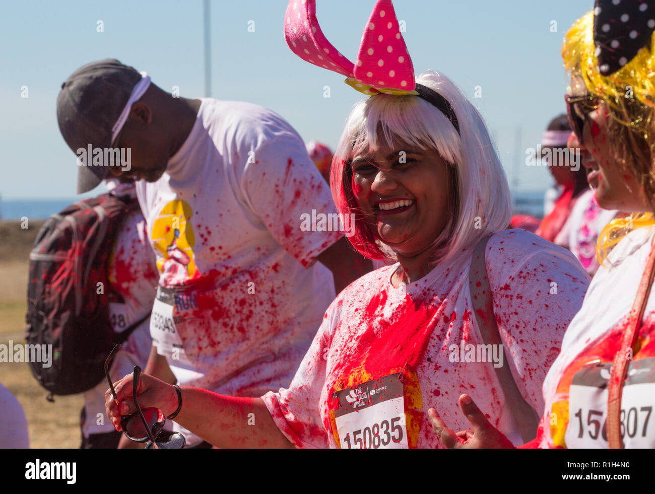 mixed race woman wearing a pair of pink polka dot bunny ears and blonde wig smiling at camera walking with group of friends in fun run sports event Stock Photo