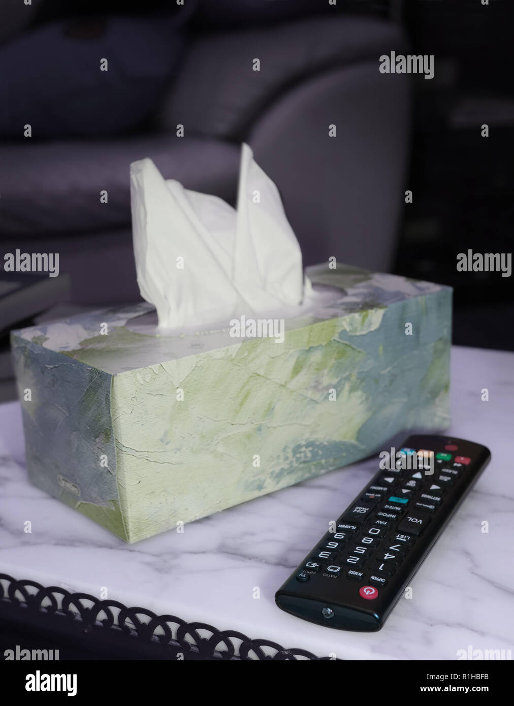 On the marble table there is a remote control and a box of tissues. The scene is illuminated by the TV screen. Stock Photo