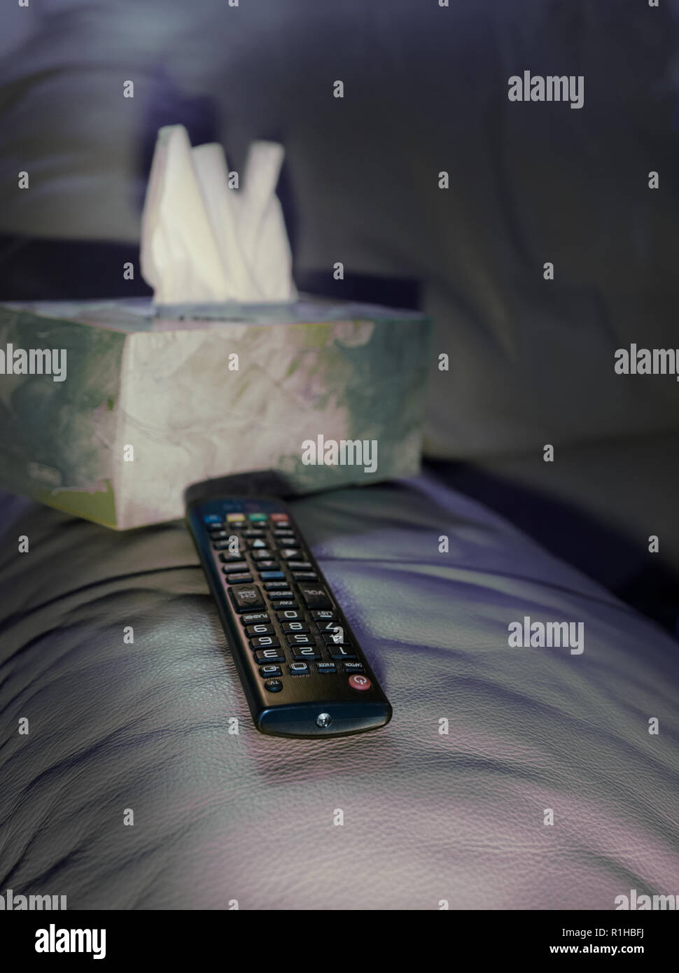 On the arm of the chair there is a remote control and a box of tissues. Reflections of the light of the tv illuminate the space. Stock Photo
