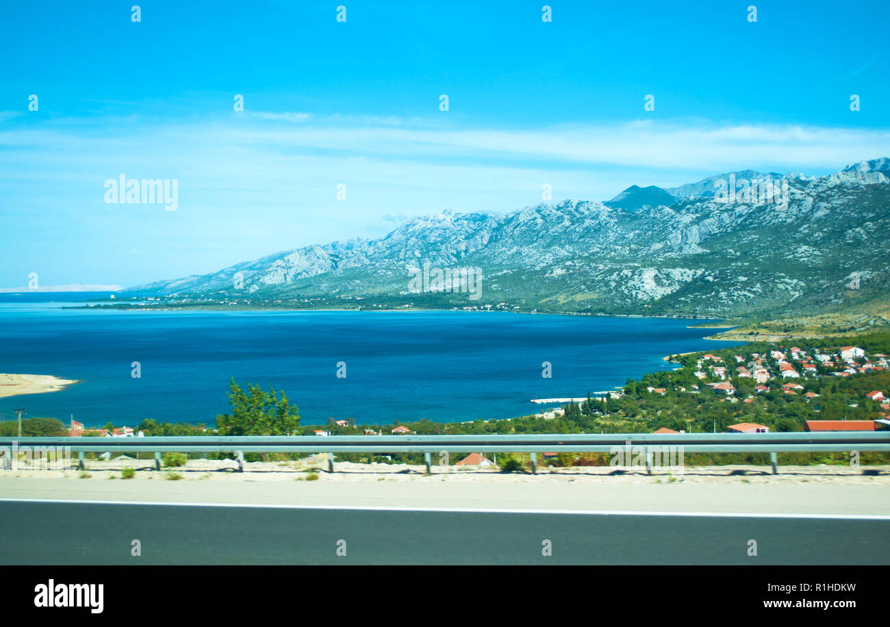 Highway on the background of Velebit mountain range, small houses among green trees and azure blue sea water on a summer day. Croatia Stock Photo