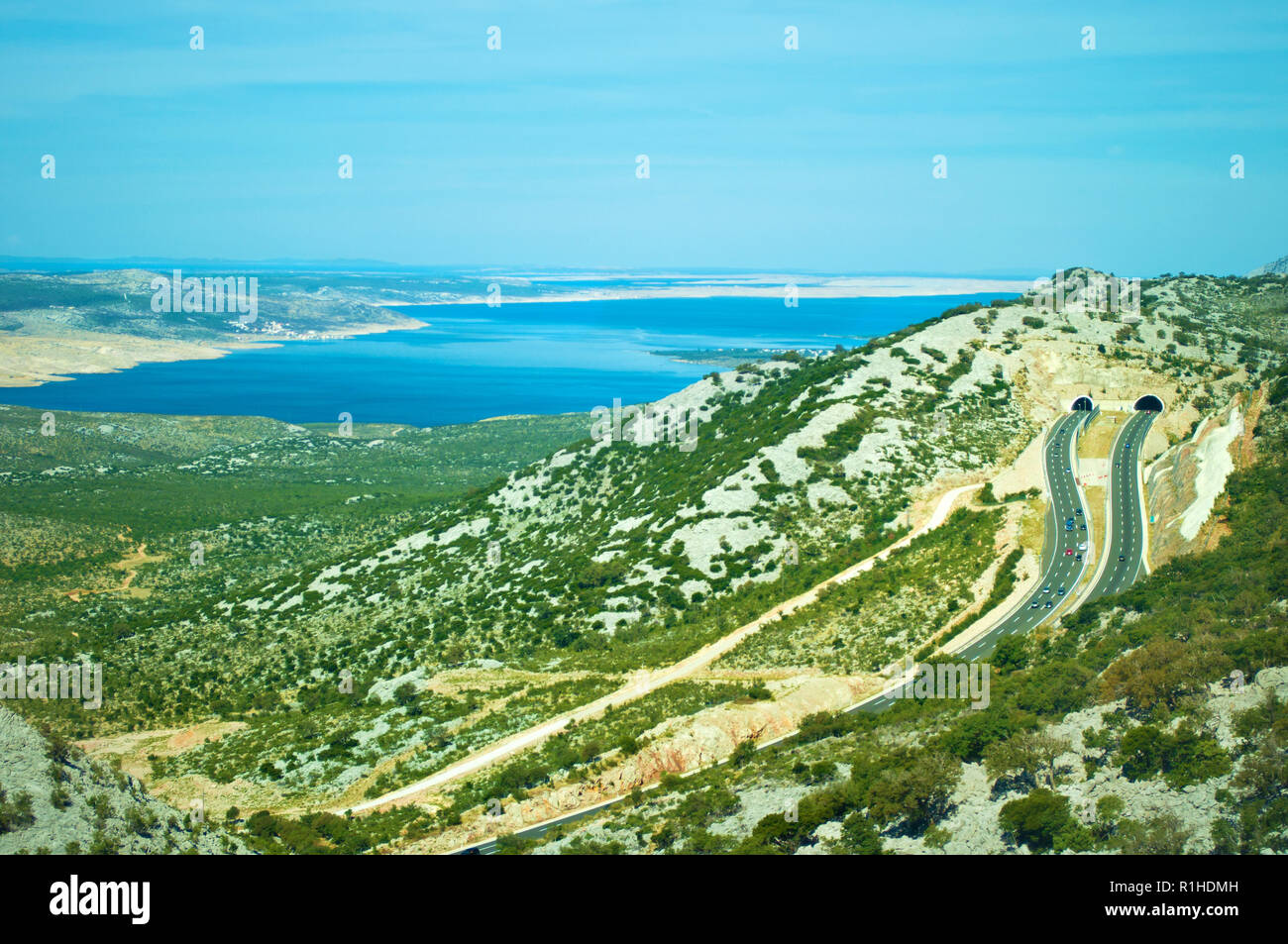 Two roads on a highway leading to tunnels in the Velebit mountain range among white rocks, green bushes and trees and azure blue sea water. Cloudless  Stock Photo
