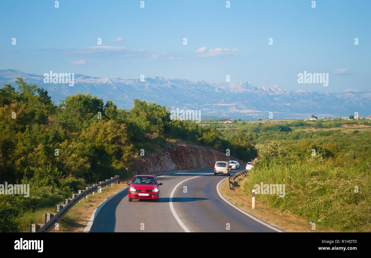 One red car on a highway in Croatia against the mountain range, many green trees and little houses on a warm summer day. Blue sky on the back Stock Photo