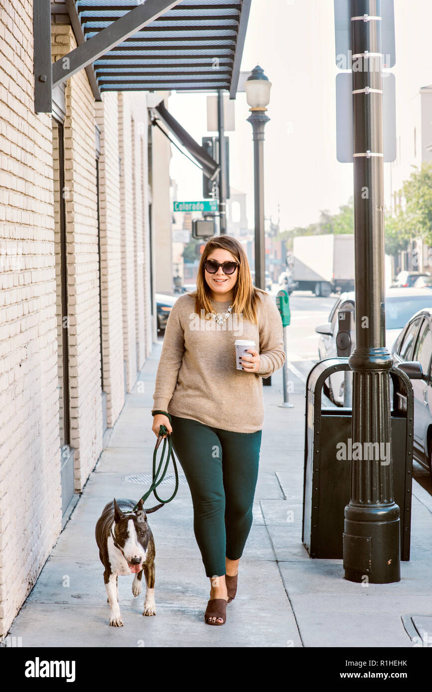 A woman walks her bull terrier dog in an urban setting while drinking coffee Stock Photo