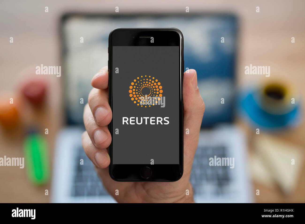 A man looks at his iPhone which displays the Reuters logo, while sat at his computer desk (Editorial use only). Stock Photo