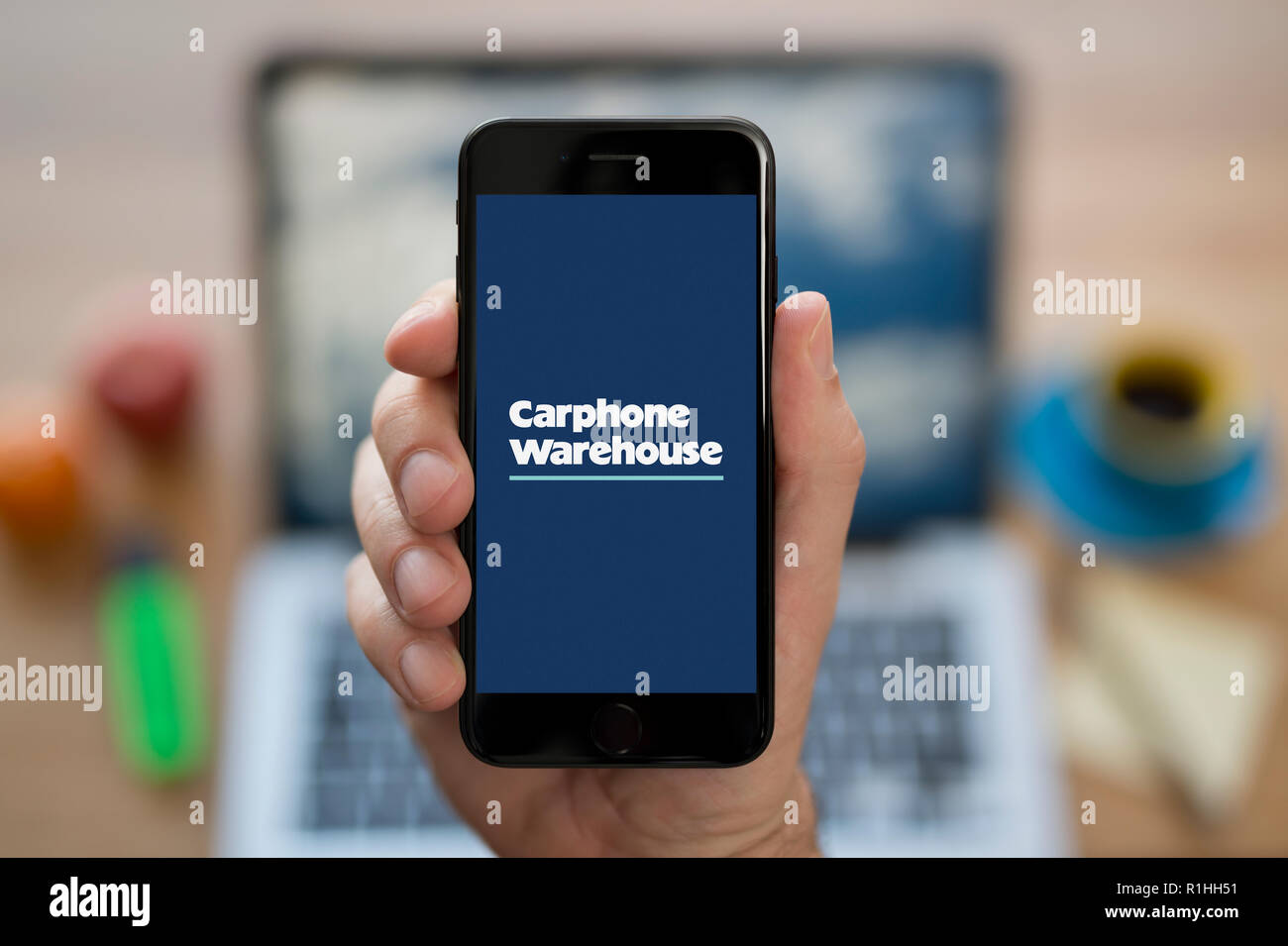 A man looks at his iPhone which displays the Carphone Warehouse logo, while sat at his computer desk (Editorial use only). Stock Photo