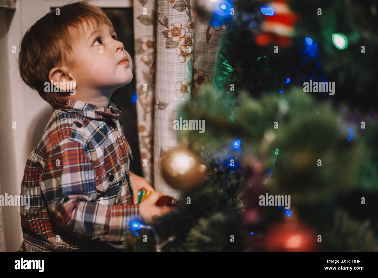 Baby boy looking at Christmas ornaments while standing beside Christmas tree at home Stock Photo