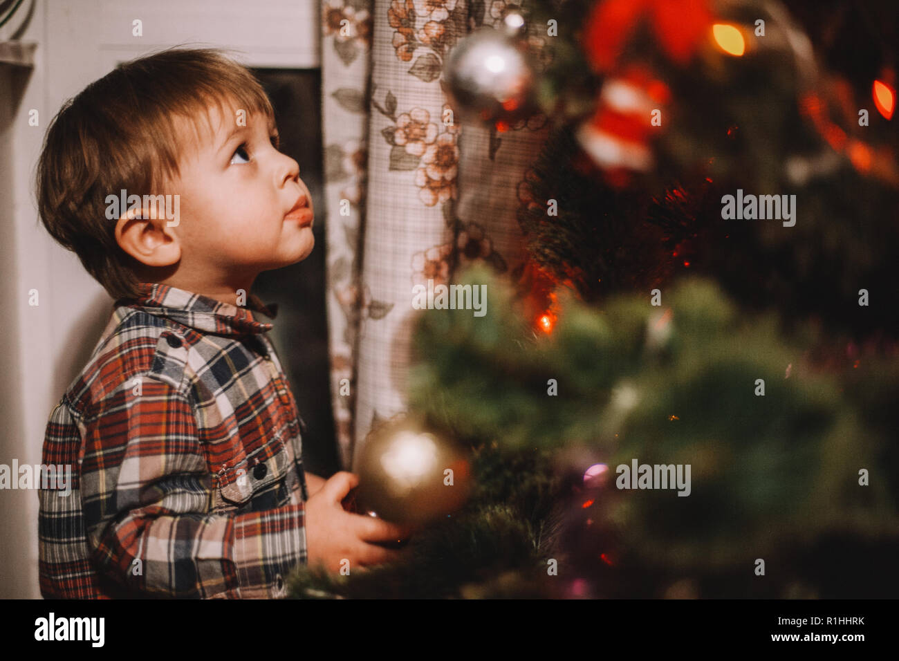 Baby boy looking at Christmas ornaments while standing beside Christmas tree at home Stock Photo
