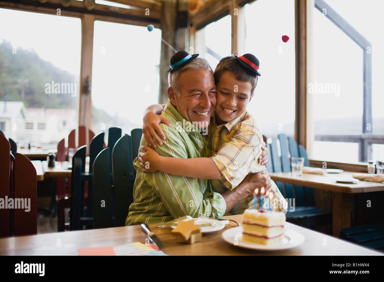 Smiling young boy and his father hugging next to a birthday cake while wearing funny hats inside a diner. Stock Photo