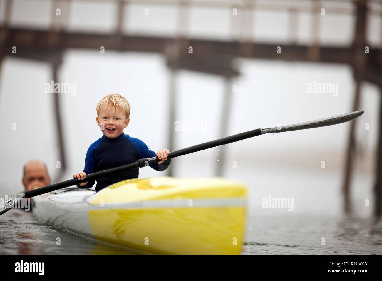 Young boy smiles as he holds an oar and sits in a kayak as a man pushes it along in the waters of a foggy harbour. Stock Photo