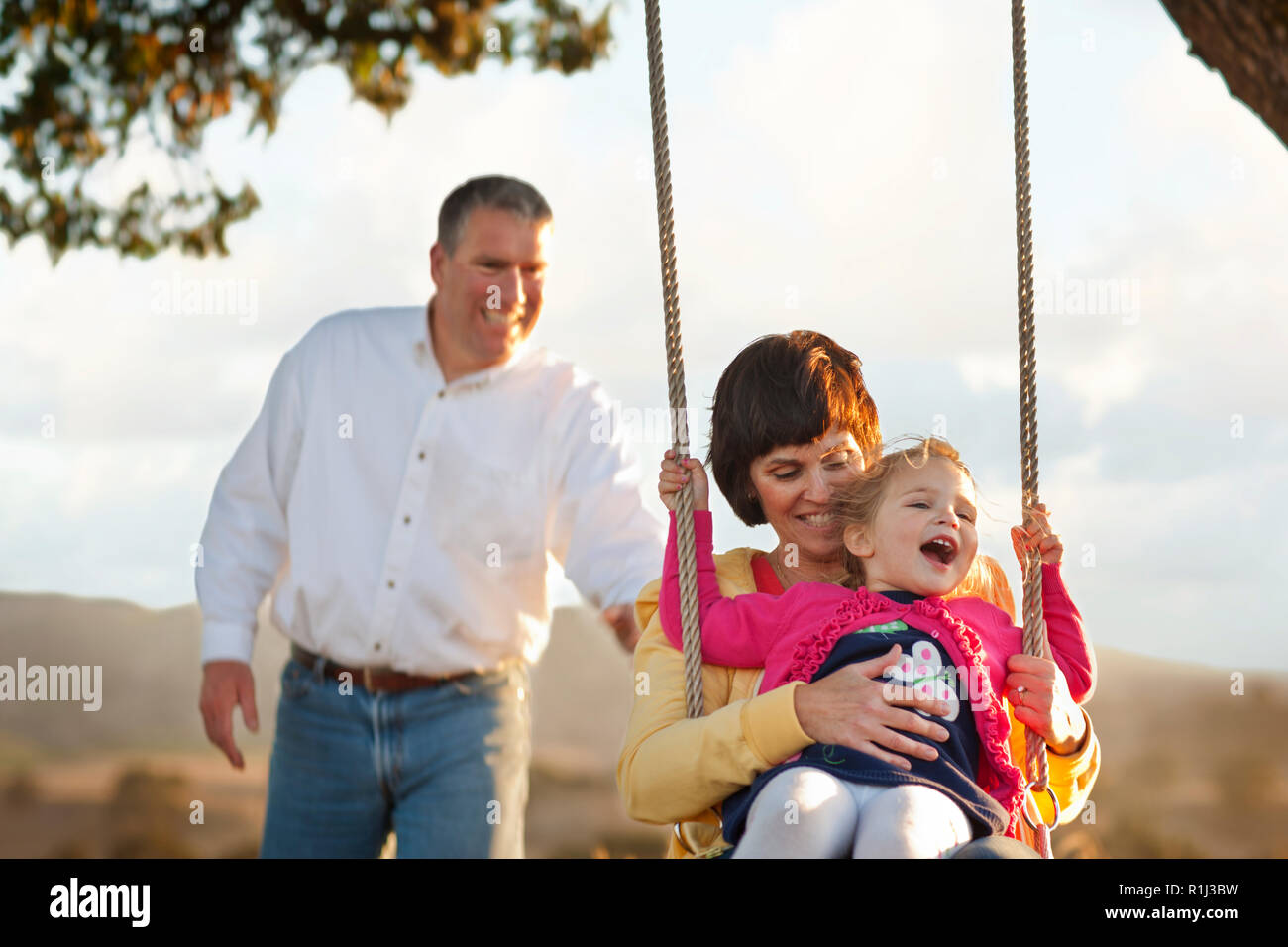 Parents Pushing Their Young Daughter On Tree Swing Stock Photo Alamy