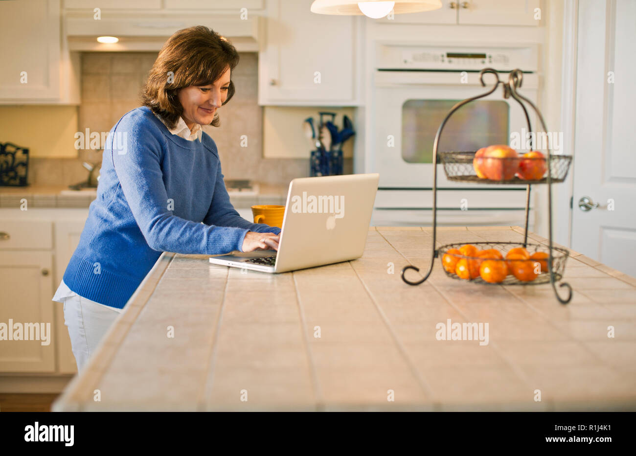 Happy mature woman typing on a laptop in her kitchen. Stock Photo
