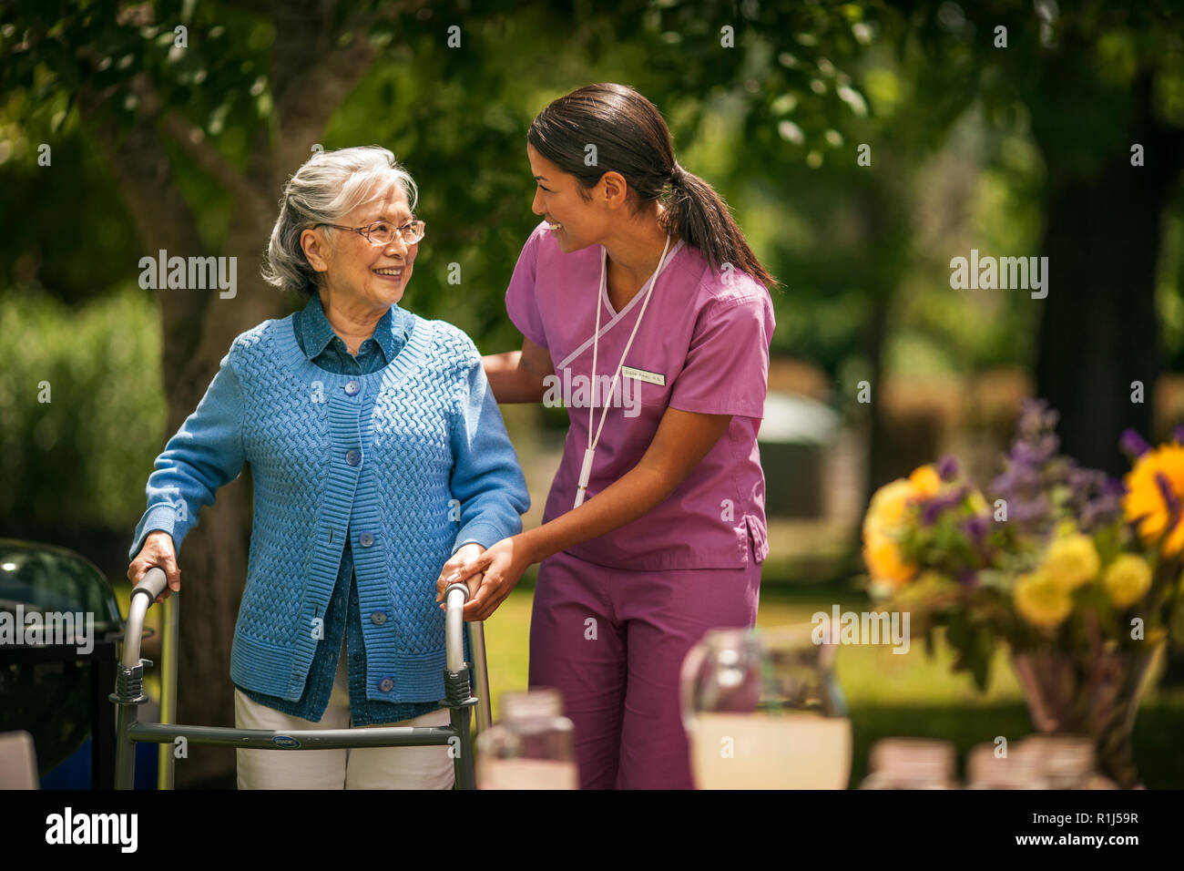 Friendly young nurse helps an elderly lady exercise with her walker in a sunny garden. Stock Photo