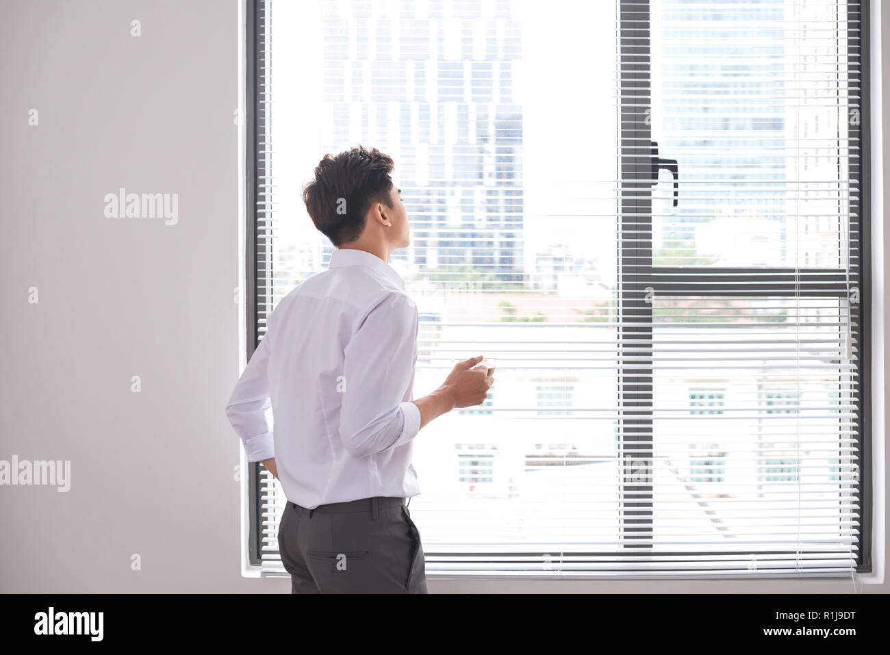 Portrait of a confident young businessman holding a cup of coffee while standing near office window, intelligent men in white shirt while resting afte Stock Photo