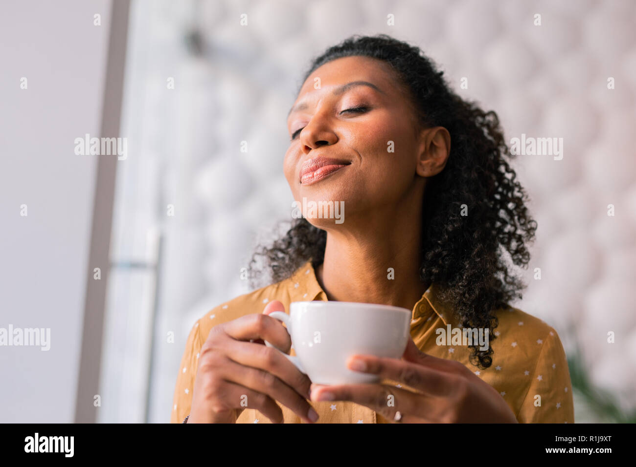 Curly dark-haired appealing woman feeling joyful while drinking coffee Stock Photo