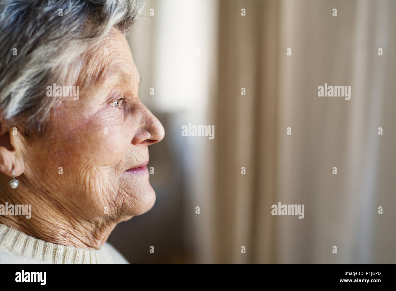 A close-up portrait of a senior woman at home, looking out of a window. Stock Photo