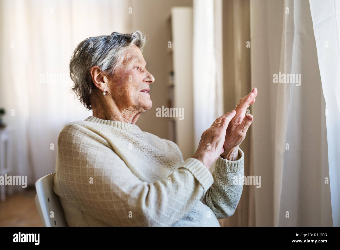 A portrait of a senior woman sitting at home, looking out of a window. Stock Photo