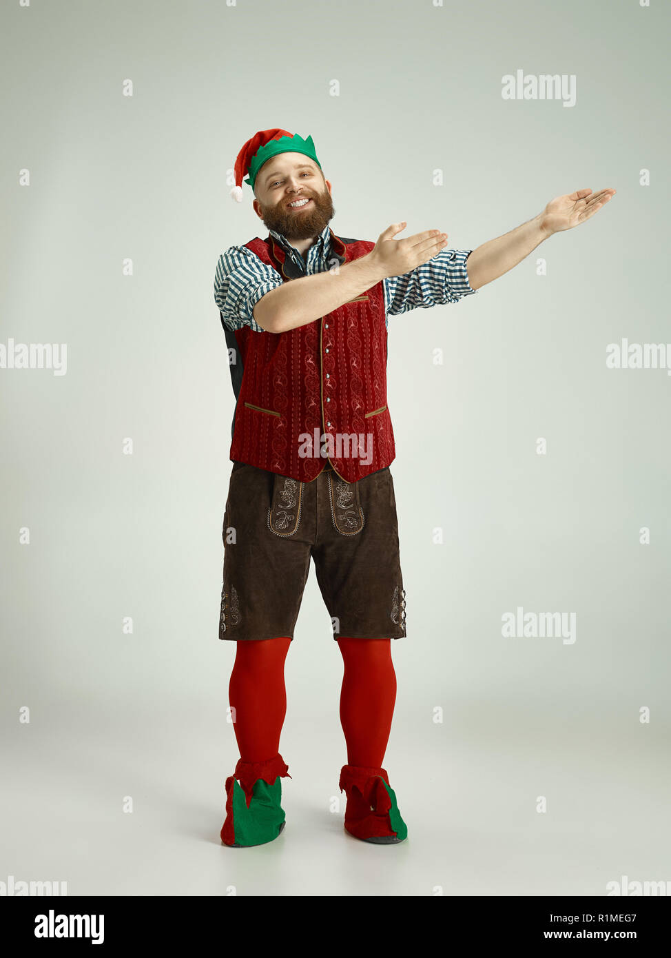 The happy smiling friendly man dressed like a funny gnome or elf pointing to left on an isolated gray studio background. The winter, holiday, christmas concept Stock Photo