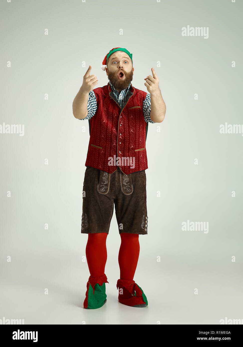 The happy smiling friendly man dressed like a funny gnome or elf pointing to camera on an isolated gray studio background. The winter, holiday, christmas concept Stock Photo