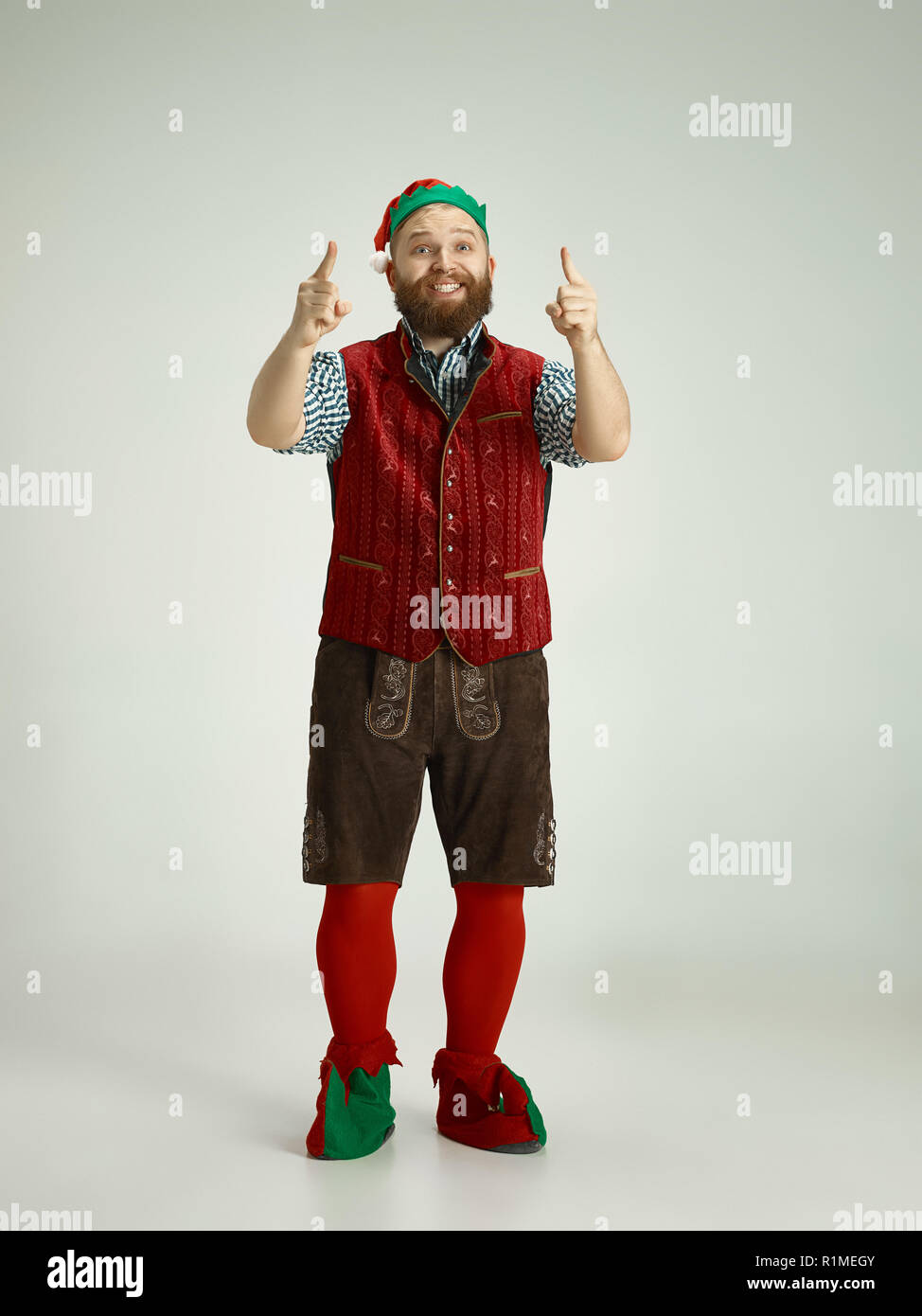 The happy smiling friendly man dressed like a funny gnome or elf pointing up on an isolated gray studio background. The winter, holiday, christmas concept Stock Photo