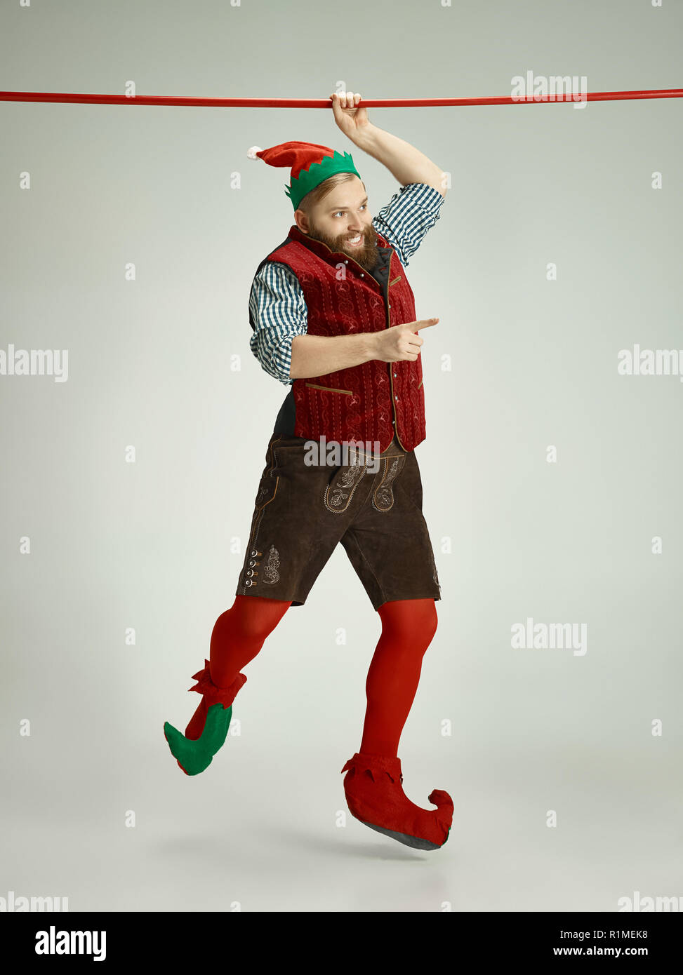 The happy smiling friendly man dressed like a funny gnome or elf hanging on an isolated gray studio background. The winter, holiday, christmas concept Stock Photo