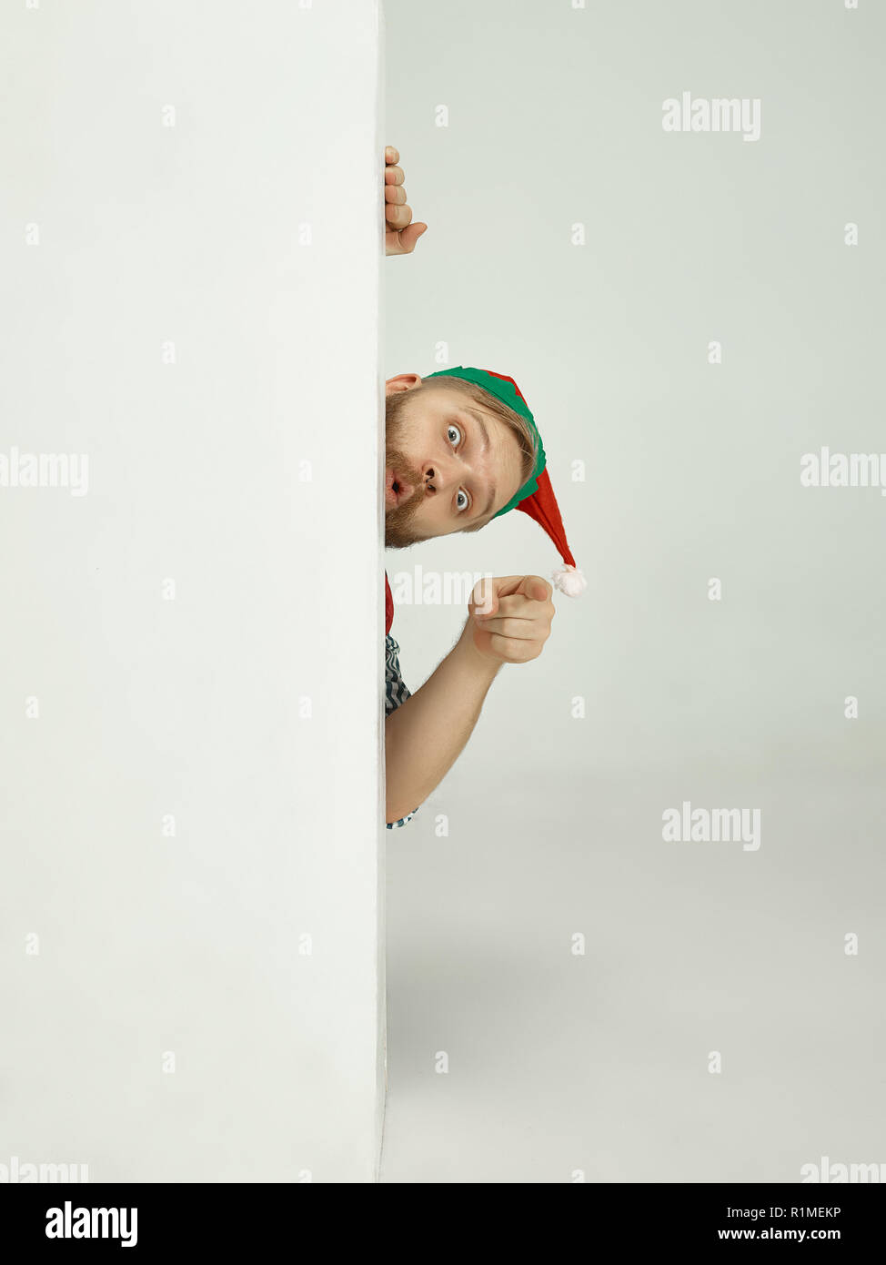 The happy smiling friendly man dressed like a funny gnome or elf pointing to camera on an isolated gray studio background. The winter, holiday, christmas concept Stock Photo