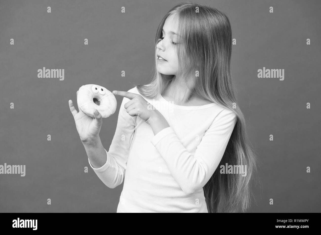 Girl holding fluffy doughnut. Glazed bagel with eyes, creative dessert. Kid eating icing out off doughnut, sweet tooth heaven. Child with long hair wearing white jumper isolated on purple background. Stock Photo