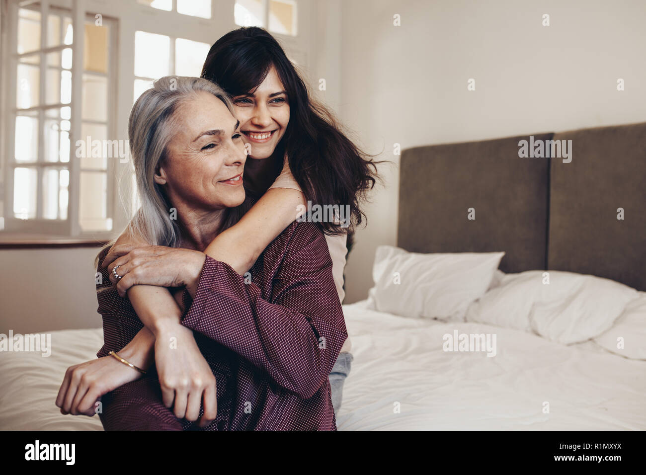 Elderly women sitting on bed with her daughter holding her hands. Smiling mother and daughter sitting at home spending time together. Stock Photo