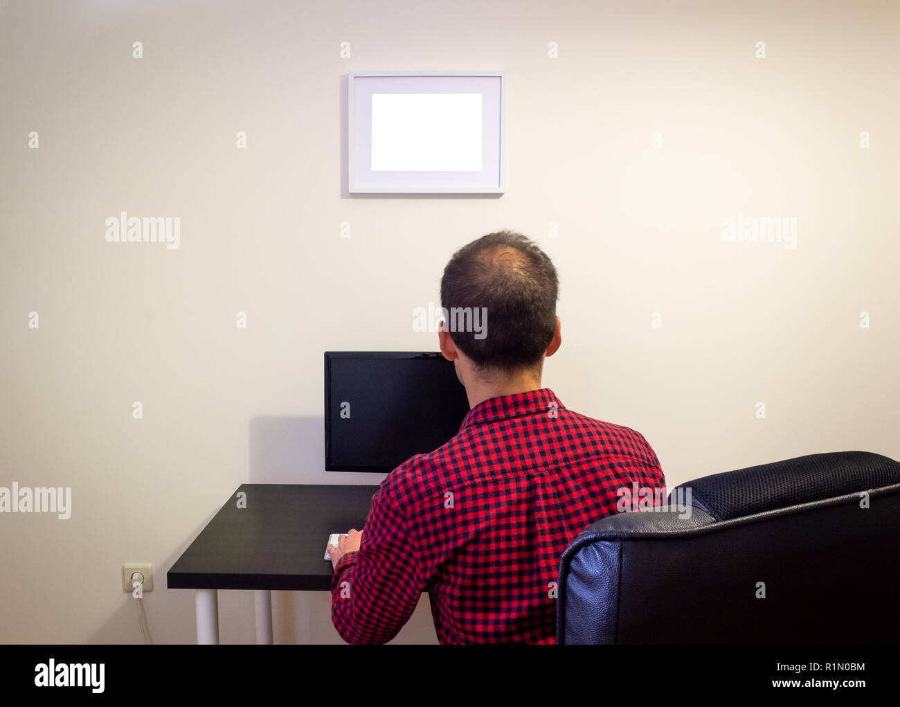 Man at Office Computer on Wooden Black Desk Mockup. Dotted Red Shirt, LCD Screen, Keyboard, Mouse, Desktop Computer, Photo Frame, Office Chair. Back V Stock Photo