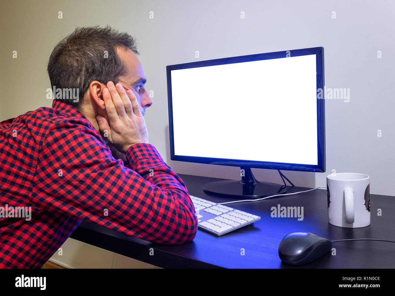 Surprised Man Stares at Office Computer on Wooden Black Desk Mockup. Dotted Red Shirt, LCD Screen, Keyboard, Mouse, White Mug. Copy Space. Stock Photo