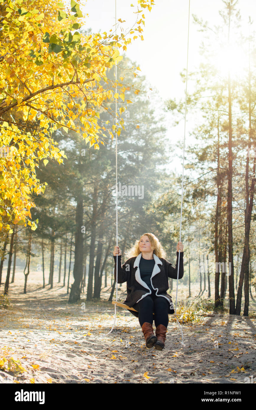 Young caucasian blonde woman in brown cardigan riding a swing in yellow autumn forest. Vertical orientation. Stock Photo